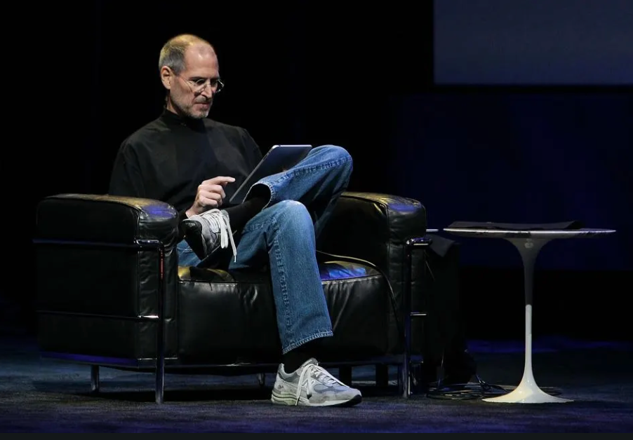 Steve Jobs sits in a large comfy-looking chair with his legs crossed as he demos the iPad